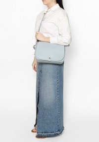 Borsa a tracolla in pelle azzurro chiaro con chiusura a patta, superficie texturizzata e tracolla regolabile. Indossata con una camicia bianca e una gonna di jeans.