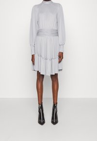 Robe gris clair avec des manches longues, un col montant, une taille froncée et un ourlet à volants. Associée à des bottines noires à la cheville avec une fermeture éclair avant.