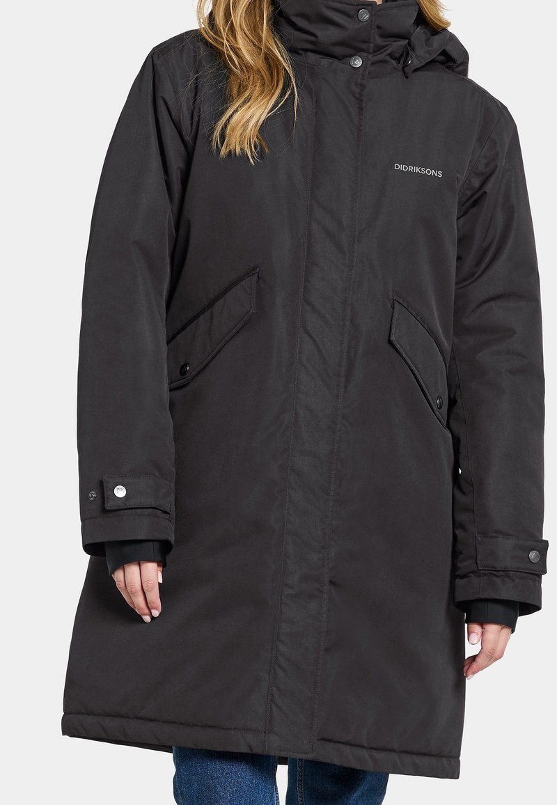 Manteau d'hiver long noir avec col montant, poches à rabat boutonnées et poignets boutonnés, porté avec un jean bleu.