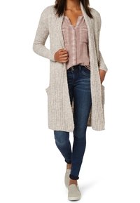 Long beige knitted cardigan with ribbed texture, open front, and side pockets, worn over a pale pink blouse and dark blue jeans.