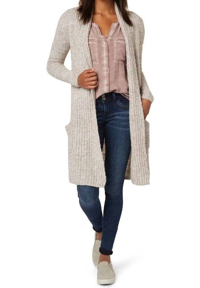 Long beige knitted cardigan with ribbed texture, open front, and side pockets, worn over a pale pink blouse and dark blue jeans.