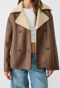 Veste en shearling brun à double boutonnage avec un grand col crème, portée sur un haut cropped en maille blanche et un jean bleu.