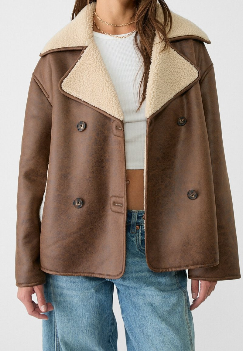 Veste en shearling brun à double boutonnage avec un grand col crème, portée sur un haut cropped en maille blanche et un jean bleu.