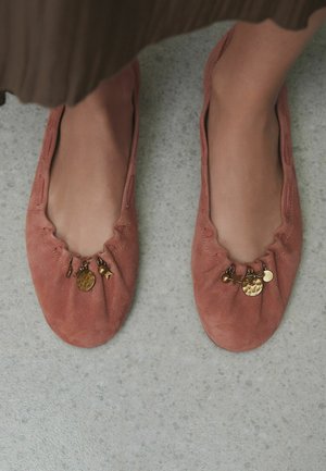 Weiche rosa Wildleder-Ballerinas, jeweils mit kleinen goldenen Anhängern verziert, darunter Scheiben, ein Stern und ein Kreuz, auf einem grauen strukturierten Boden.