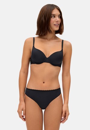 Mujer con cabello castaño hasta los hombros, vistiendo un conjunto a juego de sujetador y ropa interior negros, mirando hacia su derecha contra un fondo claro y sencillo.