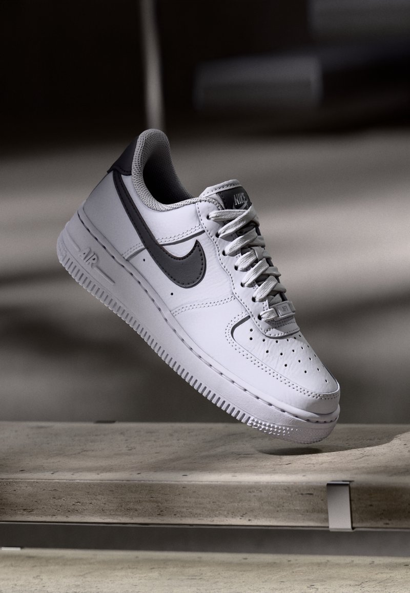 Baskets Nike Air Force 1 blanches et grises suspendues au-dessus d'une surface en béton avec des ombres douces en arrière-plan.