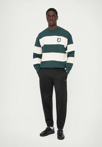 Green and cream striped sweatshirt with a logo on the chest, paired with black sweatpants and black sneakers.
