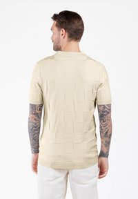 Uomo con tatuaggi su entrambe le braccia, vestito con una camicia beige a maniche corte in tessuto leggero e pantaloni bianchi, in piedi di fronte a uno sfondo bianco.