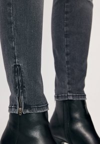 Šedé skinny džíny z denimu mají zip na kotníku a jsou doplněny černými kotníkovými botami. Materiál má mírný lesk a hladkou strukturu.