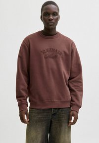Brown sweatshirt with a crew neckline, featuring an embossed "ORIGINALS" logo. The fabric appears soft, with ribbed cuffs and hem.