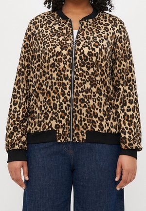 Kvinde iført en leopardmønstret lynlåstrøje med sorte manchetter og kant, kombineret med mørkeblå jeans, stående mod en neutral baggrund.