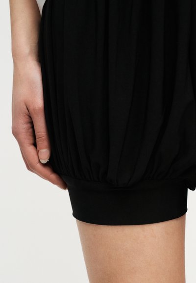 Χέρι που ακουμπά δίπλα σε μαύρη φουστίτσα με ρυτιδώσεις, που τελειώνει πάνω από το γόνατο με εφαρμοστή ζώνη στο τελείωμα, πάνω σε απλό ανοιχτόφωτο φόντο.