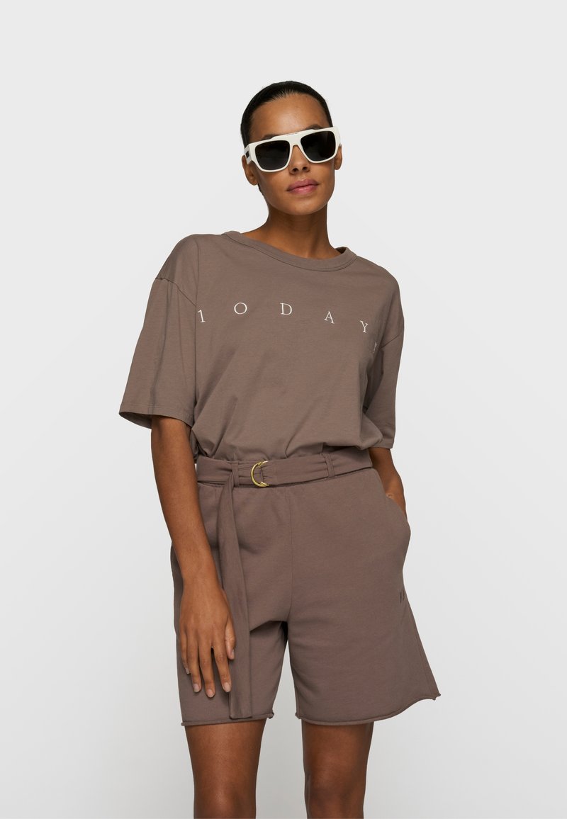 Femme portant un t-shirt taupe oversize avec le texte "1 O D A Y", un short assorti ceinturé et de grandes lunettes de soleil blanches, debout devant un fond uni.