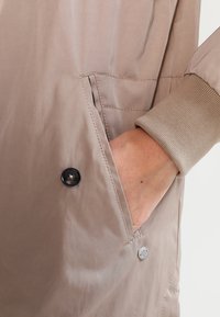 Chaqueta beige clara con una textura suave, un bolsillo lateral con un botón negro y un puño acanalado en un tono beige complementario.