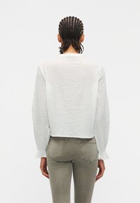 Άτομο με πλεγμένα μαλλιά που φοράει λευκό μπλουζάκι με μακριά μανίκια και υφή, καθώς και εφαρμοστά χακί τζιν, εμφανιζόμενο από πίσω σε απλο φόντο.