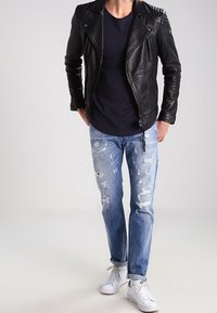 Musta nahkainen pyöräilijän takki, jossa vetoketju ja tikatut yksityiskohdat, yhdistetty vaaleanpesuihin revittyihin farkkuihin ja valkoisiin tennareihin.