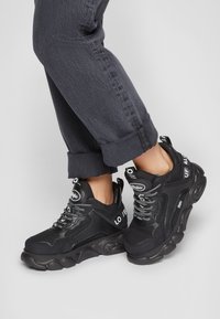 Personne portant des baskets noires volumineuses avec la marque blanche "Buffalo" et un pantalon noir retroussé sur un fond clair.