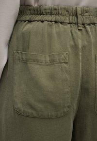 Κοντινό πλάνο ελιάς πράσινου παντελονιού με ελαστική ζώνη μέσης και μια μεγάλη πίσω τσέπη ραμμένη στη δεξιά πλευρά.