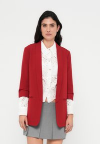 Κόκκινο σακάκι με δομημένο σχέδιο, που διαθέτει μακριά μανίκια και εμπρόσθιες τσέπες, συνδυασμένο με λευκό πουκάμισο με διάτρητα σχέδια και γκρι πλισέ φούστα.