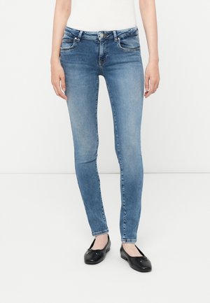 Jean skinny bleu en denim avec une texture délavée, cinq poches et une taille avec passants de ceinture, associés à des chaussures plates noires.