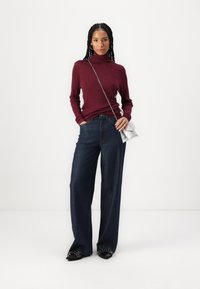 Suéter de cuello alto burdeos combinado con jeans de pierna ancha azul oscuro. La modelo lleva un bolso cruzado plateado y botas de tobillo negras con detalles.