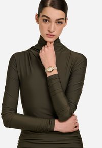 Olivno zelena majica s dugim rukavima i visokim ovratnikom te naborima. Model nosi zlatno-bež sat na glatkoj narukvici.