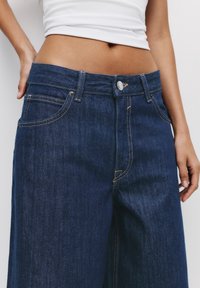 Jean en denim bleu foncé à taille haute, avec une fermeture à boutons et zip à l'avant, cinq poches et des surpiqûres jaunes contrastées.