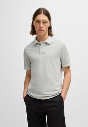 Lichtgrijze polo shirt van katoen met een knoopsluiting van drie knopen, korte mouwen en geborduurd logo op de borst. Zwarte broek casual gedragen.