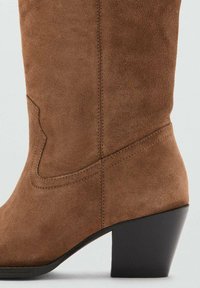Bota vaquera de antelina marrón con un diseño afinado, punta puntiaguda y tacón negro grueso. Presenta detalles acolchados y una ligera curva en el caña.