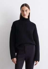 Schwarzer Strickpullover mit hohem Kragen, geripptem Muster und lockerer Passform. Lange Ärmel und leicht verkürzter Saum. Kombiniert mit dunklen, schmalen Hosen.