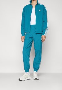 Tuta color turchese con accenti bianchi a tre strisce. Giacca con zip e pantaloni con vita elastica in materiale leggero, abbinati a sneakers bianche.