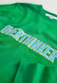 Zelené bavlnené tričko s vyšívaným nápisom "OVERTHINKER" v modrej farbe so žltým obrysom, s hladkou textúrou a dizajnom s okrúhlym výstrihom.