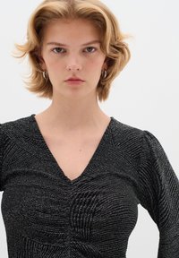 Top nero testurizzato con scollatura a V, tessuto morbido con fili metallici che creano un motivo ondulato e maniche lunghe con leggere arricciature.