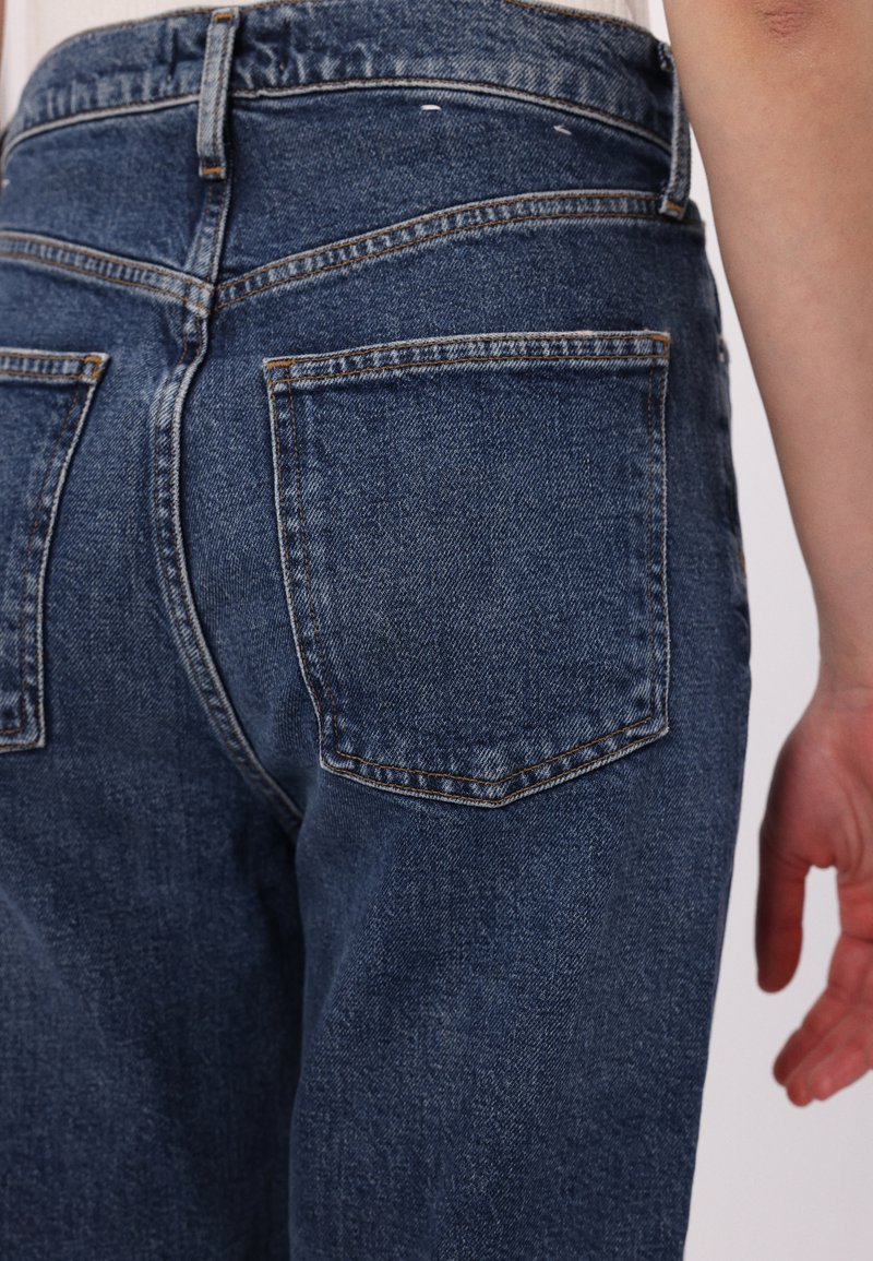 Jeans en denim bleu foncé à taille haute, dotés de poches arrière et de coutures contrastées. La texture est lisse avec une coupe classique.