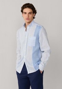 Giovane con capelli castani mossi che indossa una camicia a righe verticali blu e bianche con bottoni e pantaloni blu navy, mani nelle tasche, sfondo neutro.