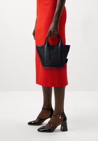 Borsa tote in pelle nera con manici corti, caratterizzata da un design geometrico. Texture liscia con un colore solido. Indossata con un vestito rosso e tacchi neri.