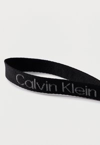 Μαύρο υφαντό υφαντό βραχιόλι με το κέντημα "Calvin Klein" σε γκρι νήμα, απεικονίζεται σε απλό λευκό φόντο.