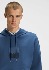 Felpa blu realizzata in tessuto morbido, caratterizzata da un grande logo quadrato nero con "HUGO" al centro, completata da dettagli con lacci.