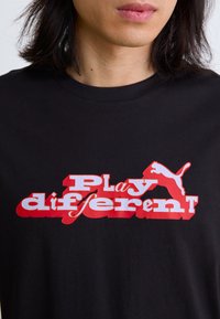 Sort t-shirt i sort bomuld med fed rød og hvid tekst "Play different" i kontrasterende skrifttyper og et rødt Puma-logo.