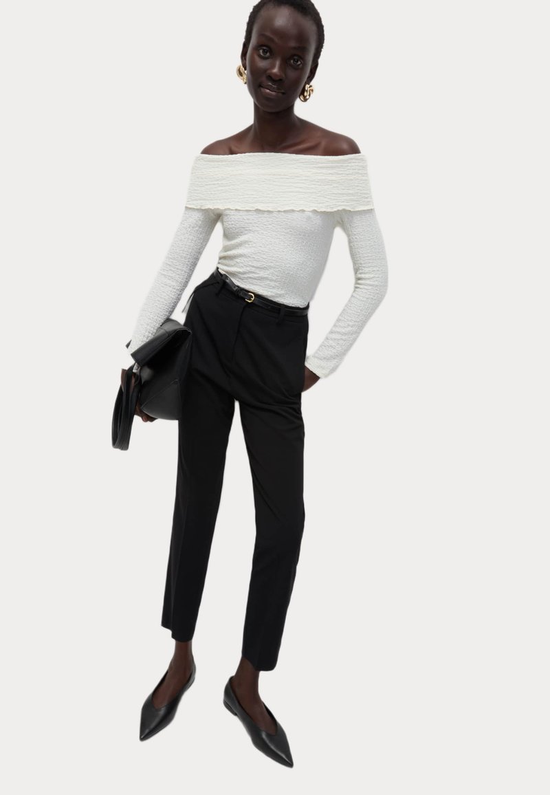 Witte off-the-shoulder top van gestructureerde stof, in combinatie met zwarte high-waisted broek en zwarte puntige flats, met een zwarte tas.