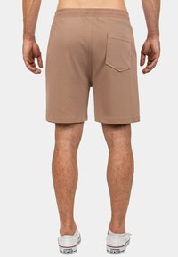 Pantaloni corti marroni con vita elastica e tasca posteriore, caratterizzati da una texture liscia e un taglio dritto, abbinati a sneakers bianche.