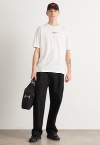 Hvid T-shirt med broderet logo, sorte cargo-bukser, sort kasket med rød accent, holder en rektangulær sort taske, blanke sorte sko.
