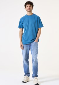 Türkisfarbiges Baumwoll-T-Shirt mit Rundhalsausschnitt und kurzen Ärmeln, kombiniert mit hellblauen Jeans und beigen Sneakern. Einfaches Design, keine sichtbaren Muster.