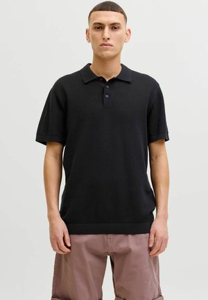 Jeune homme aux cheveux courts portant un polo en maille noir à manches courtes et un pantalon marron clair, debout devant un fond clair uni.