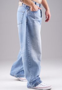 Hellblaue Jeans im lockeren Schnitt, mit zwei Gesäßtaschen, sichtbaren Nähten und einem abgenutzten Finish. Kombiniert mit weißen Sneakers.