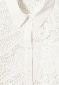 Blouse blanche à col avec des motifs complexes en dentelle et des détails en crochet floral translucide sur le devant et la fermeture à boutons.