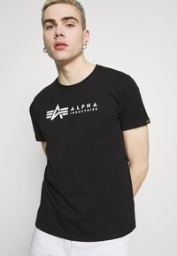Juodas medvilnės marškinėliai su baltu logotipu "ALPHA INDUSTRIES", stilizuota raide 'A' ir juostelėmis. Trumpomis rankovėmis, standartiniu apvaliu kaklu.