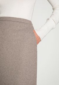 Falda marrón texturizada de corte recto, con un bolsillo lateral, combinada con una blusa blanca de mangas largas acanaladas.