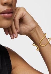 Brazalete de metal amarillo que se envuelve en una forma minimalista y fluida; textura suave con un brillo sutil. Lleva puesto en una muñeca desnuda.