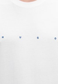 Chemise blanche avec de petites lettres bleues espacées formant le mot "HUGO" sur la poitrine.
